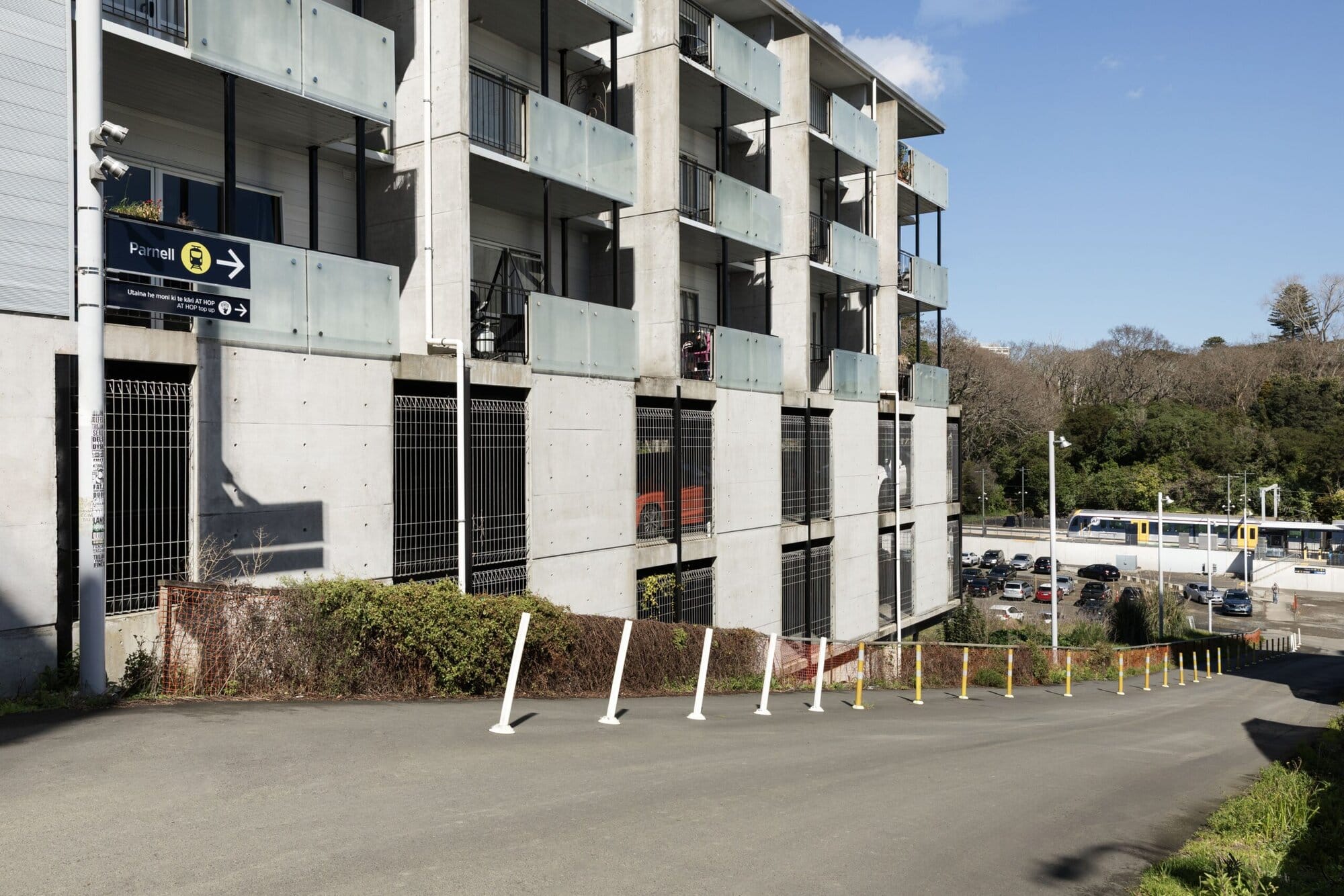 A steep, sloping roadway runs alongside a multi-storey apartment building with balconies and concrete walls. Bollards separate a pathway for pedestrians. Directional signage points toward Parnell Station.