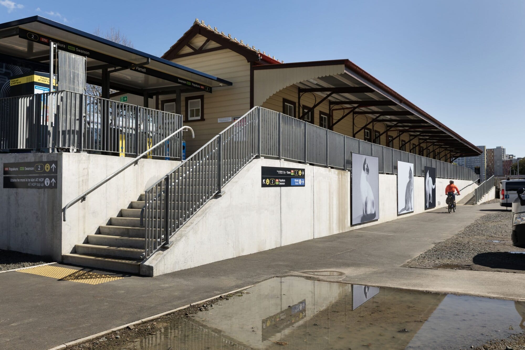 Stairs and an accessible ramp lead up to a train station platform beside a heritage-style station building. Directional signage, wall-mounted artwork, and a cyclist on the pathway are visible in the foreground.