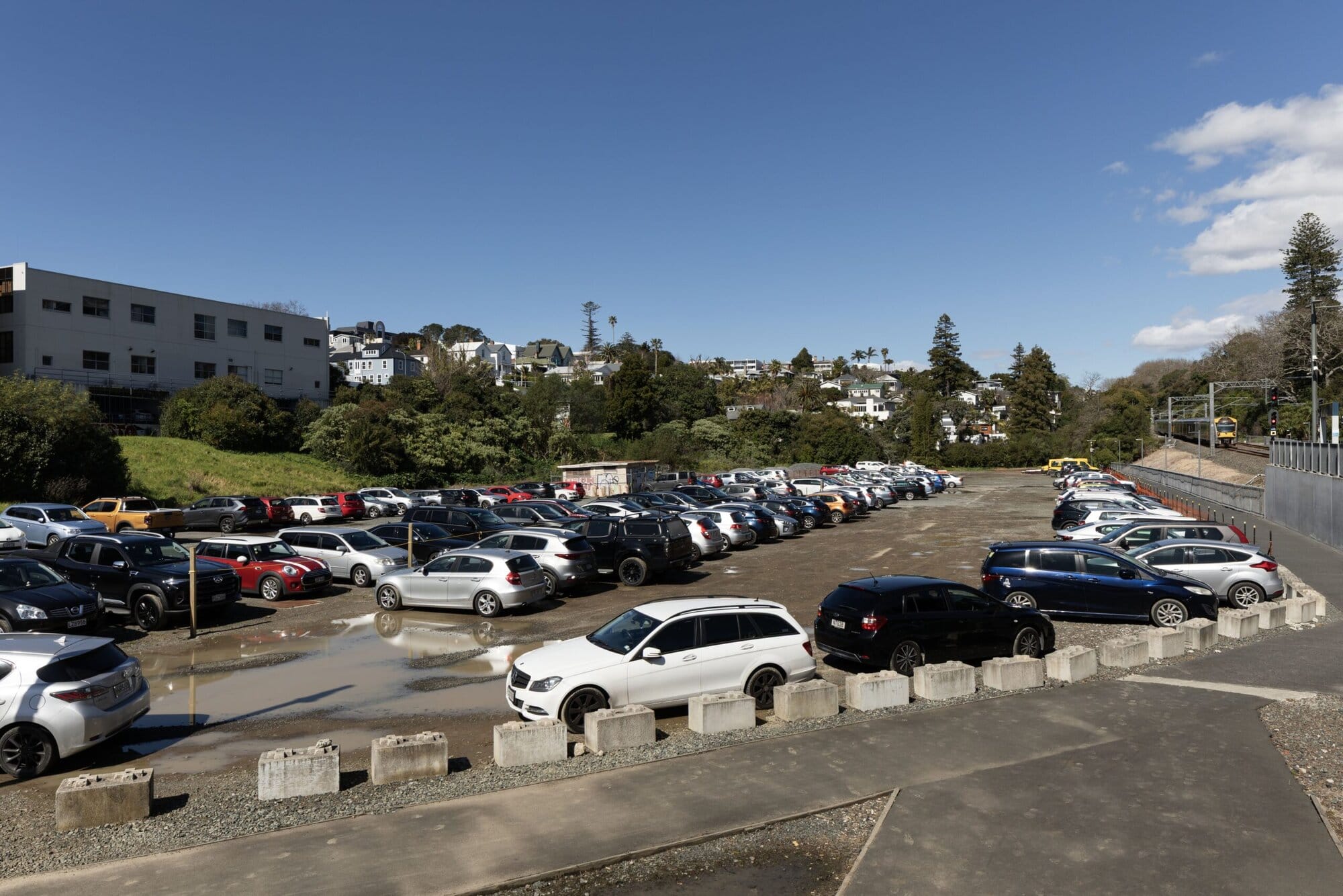 A large gravel car park with potholes and puddles sits beside a railway line. A paved pathway, separated by low concrete bollards, surrounds the car park.