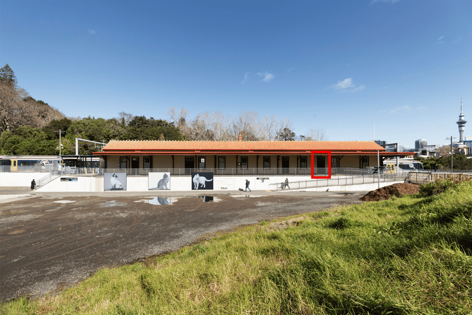 Wide view of a long, single-storey train station building with a red tiled roof and cream walls. The right-most doorway is highlighted by a red box.